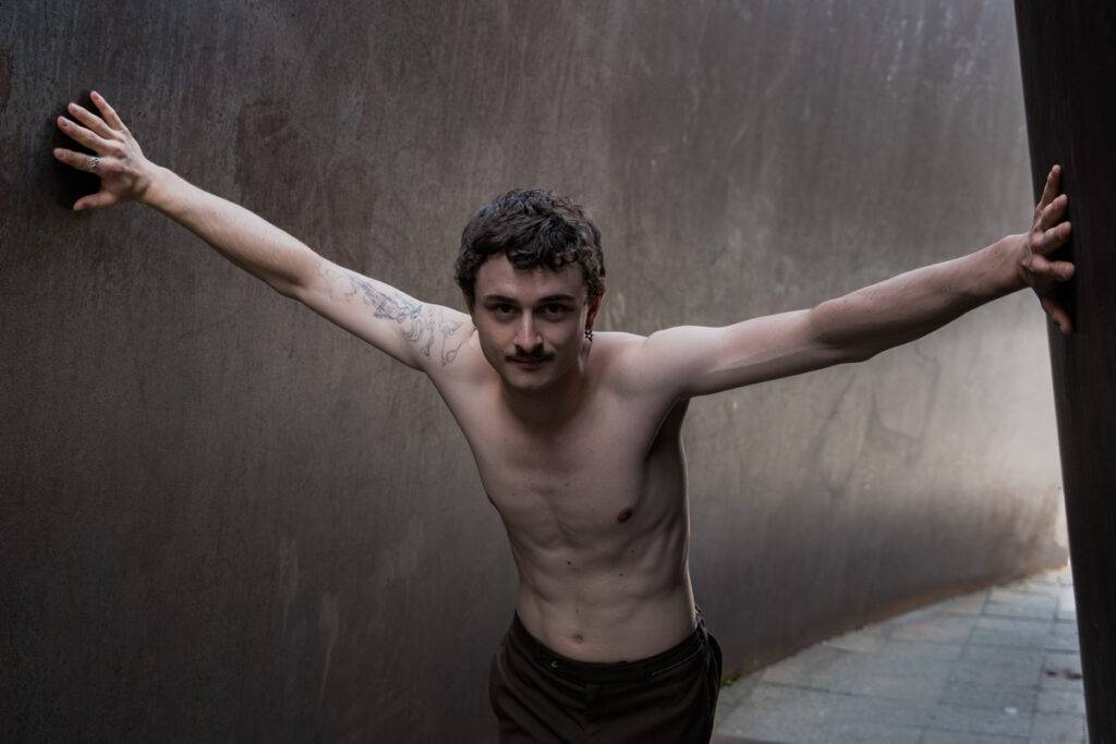 Das Bild zeigt einen Porträt-Szene eines Schauspielers, Felix Steinhilber, der vor einer grauen Metallwand mit ausgestreckten Armen steht. Die Pose vermittelt Kontemplation, Verletzlichkeit und eine gewisse Kraft. Der Hintergrund ist dunkel und unscharf, was die Aufmerksamkeit auf den Schauspieler lenkt.