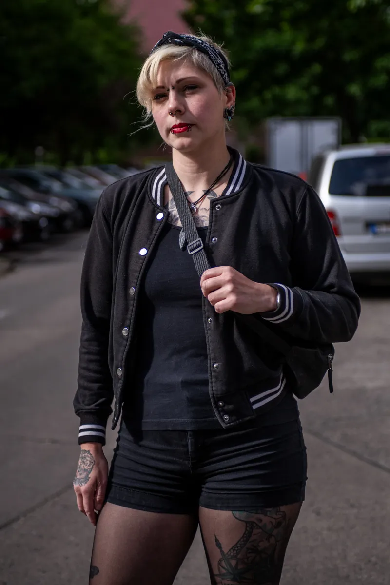  Die Aufnahme zeigt eine junge Frau, vermutlich im Alter von 25-35 Jahren, die in einer belebten Straße steht. Ihre Kleidung ist vollständig schwarz – eine schwarze Baseballjacke mit weißen Details, ein schwarzes T-Shirt, schwarze Shorts und schwarze Strumpfhosen. Ihre roten Lippen und das Bandana im Haar verleihen ihr einen rebellischen Look. Sie trägt eine schwarze Schultertasche über der Schulter. Im Hintergrund ist ein grauer Kleinwagen (wahrscheinlich ein VW Golf oder ähnliches) und ein typisches europäisches Reihenhaus zu sehen. Die Szene wirkt urban und modern.