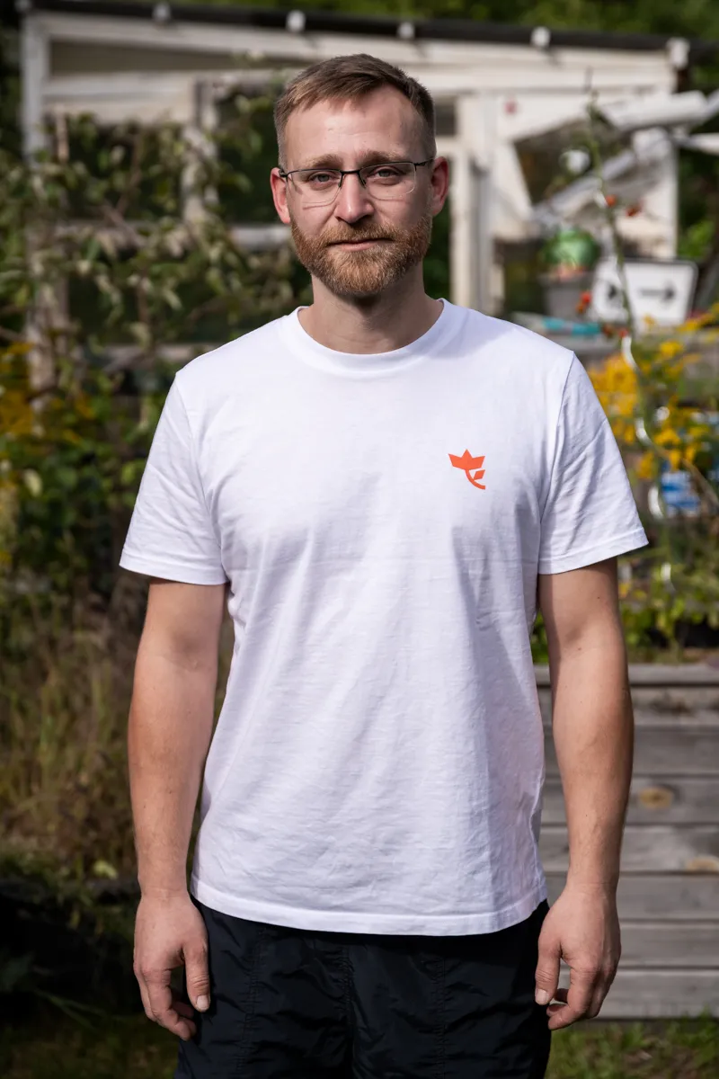 Die Aufnahme zeigt einen Mann, der vor einem Gewächshaus steht. Er trägt ein weißes T-Shirt mit einem roten Logo auf der Brust. Er trägt eine Brille und eine schwarze Hose. Im Hintergrund befindet sich ein Gewächshaus mit einem Metallgitter. Die Szene wirkt entspannt und natürlich.