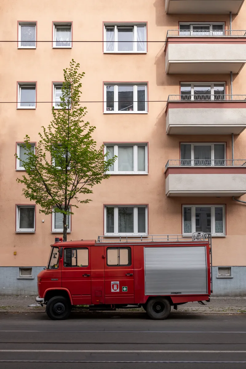 Das Bild zeigt eine straßenseitige Aufnahme eines roten Feuerwehrwagens, vermutlich ein Mercedes-Benz Unimog, der vor einem mehrgeschossigen Wohnhaus steht. Im Vordergrund steht ein Baum mit frischem, jungem Laub. Die Fassade des Wohnhauses ist in einem hellen, rosafarbenen Ton gehalten und mit vielen rechteckigen Fenstern versehen. Die Straße ist mit Stromleitungen versehen. Die Szene wirkt ruhig und urban.
