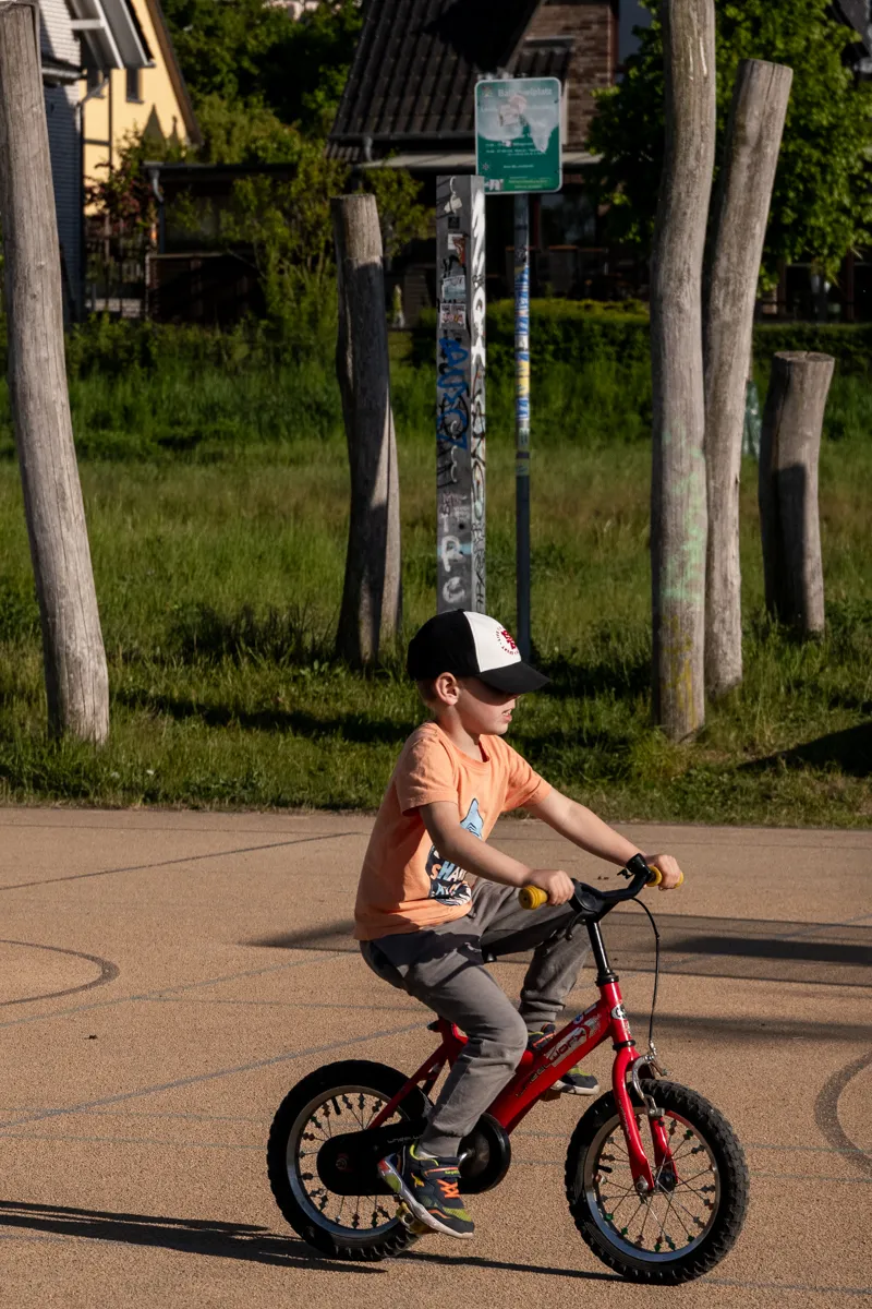 Das Bild zeigt einen jungen Mann, vermutlich im Alter von etwa 8-12 Jahren, der auf einem roten Kinderfahrrad auf einem asphaltierten Weg fährt. Er trägt einen Baseballcap, ein orangefarbenes T-Shirt mit einem Aufdruck und Jeans. Im Hintergrund befinden sich alte, verwitterte Gebäude mit Schieferdächern und Graffiti. Es sind auch Bäume und einige Holzstützen zu sehen. Der Weg ist uneben und weist Abnutzungsspuren auf. Die Lichtverhältnisse sind hell und sonnig, was auf eine warme Tageszeit hindeutet.