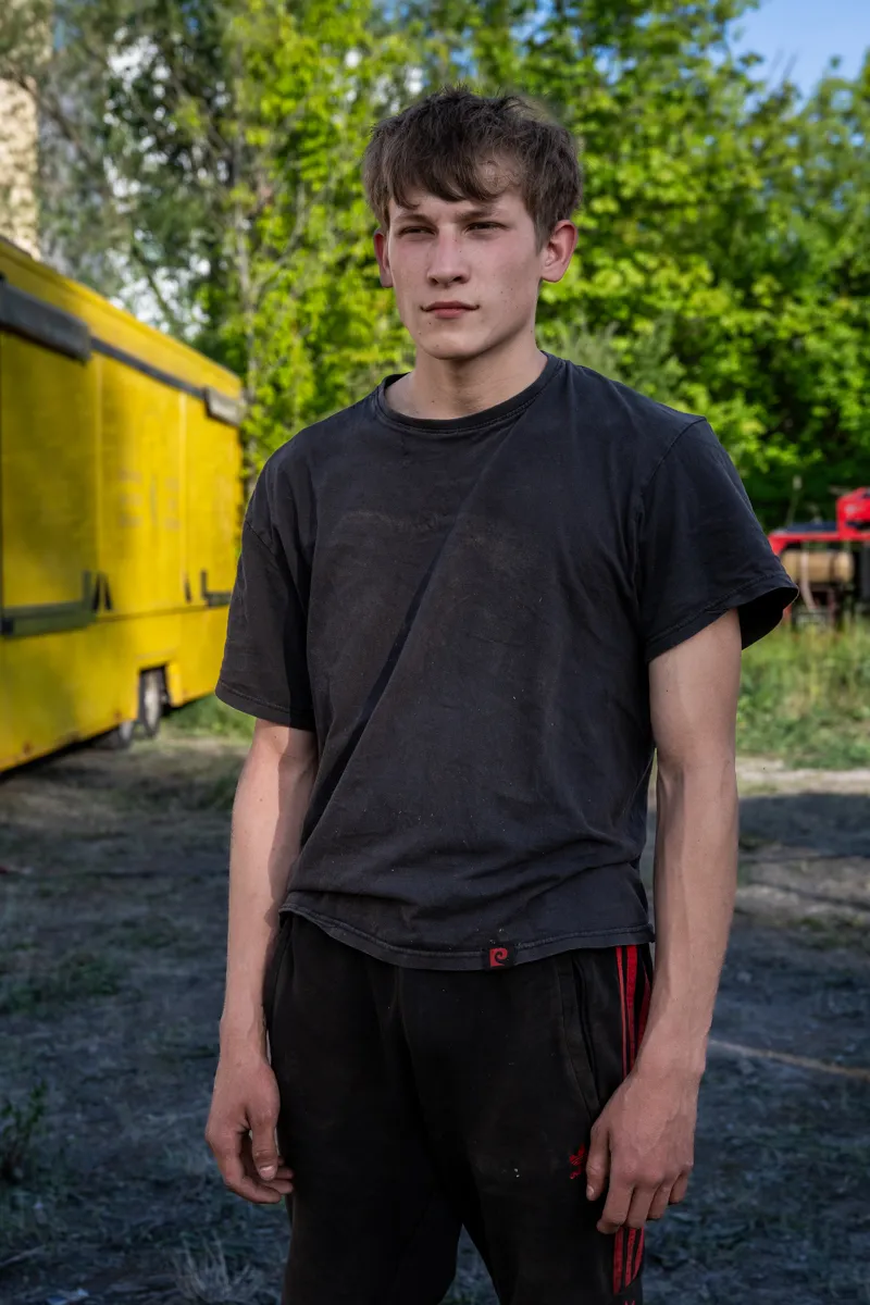 Die Aufnahme zeigt einen jungen Mann, vermutlich im Alter von etwa 20-25 Jahren, der vor einem gelben Van in einer grünen, ländlichen Umgebung steht. Er trägt ein schwarzes T-Shirt und rot-schwarze Trainingshosen. Die Szene wirkt ungestört und realistisch. Der Hintergrund ist von Bäumen und Vegetation geprägt, was auf eine ländliche oder sonstige Umgebung hindeutet.Caption: Die Aufnahme zeigt einen jungen Mann, vermutlich im Alter von etwa 20-25 Jahren, der vor einem gelben Van in einer grünen, ländlichen Umgebung steht. Er trägt ein schwarzes T-Shirt und rot-schwarze Trainingshosen. Die Szene wirkt ungestört und realistisch. Der Hintergrund ist von Bäumen und Vegetation geprägt, was auf eine ländliche oder sonstige Umgebung hindeutet.