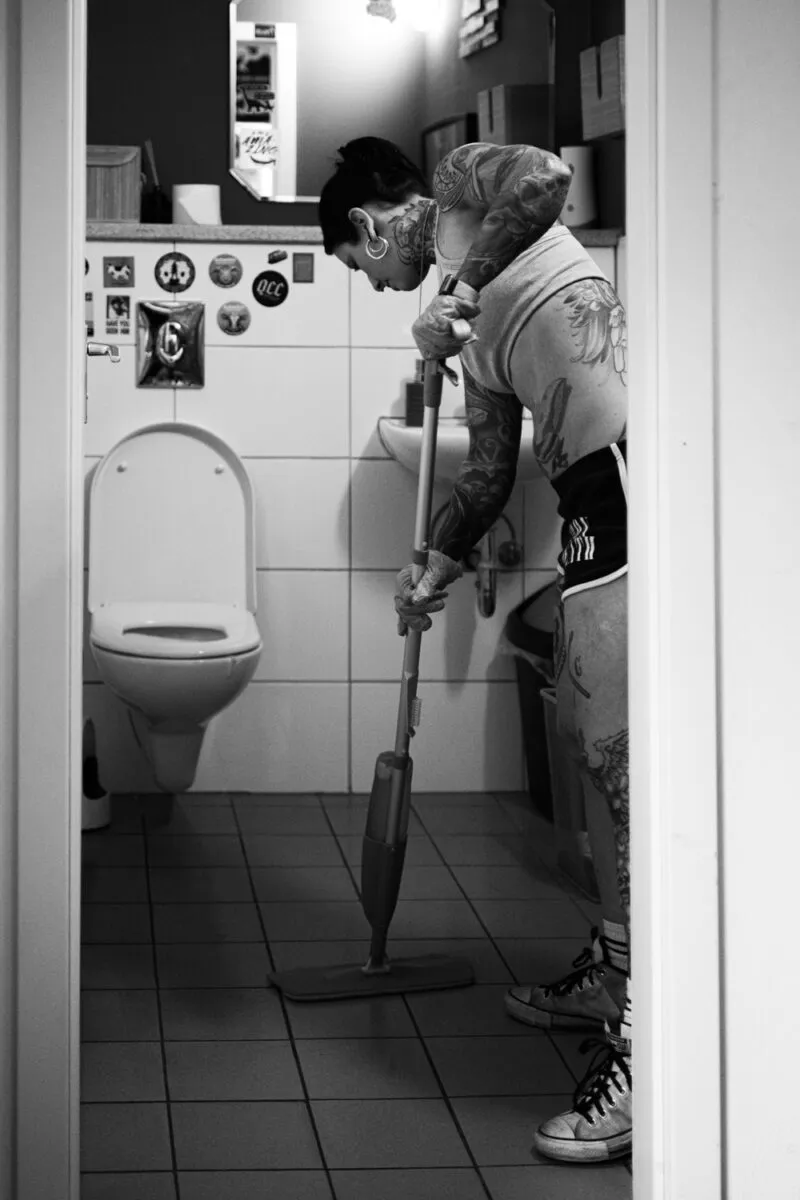 Schwarzweißes Foto einer Frau, die ein Badezimmer reinigt. Die Szene spielt sich in einem kleinen Raum ab, der zu 'Berlin Strength' gehört.