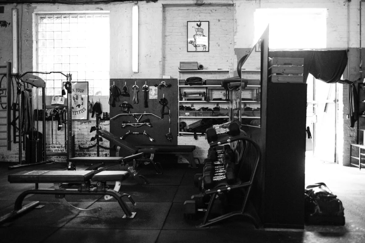 Die Schwarzweißfotografie zeigt ein Fitnessstudio, in Berlin. Im Vordergrund sind Trainingsgeräte wie eine Bankdrücker-Anlage und eine Rudermaschine zu sehen. Im Hintergrund befinden sich weitere Geräte und eine Wandregalinstallation. Eine Personal Trainerin (Caro) ist nicht direkt erkennbar, aber die Umgebung deutet auf ihre Tätigkeit hin.