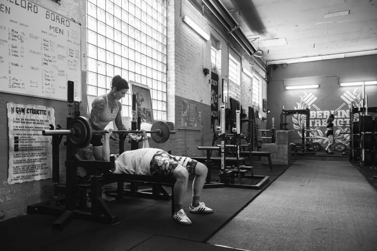 Das Schwarzweißfoto zeigt Caro, Personal Trainerin, während des Trainings im Berlin Strength Gym. Der Fokus liegt auf dem Muskelaufbau und der sportlichen Aktivität.