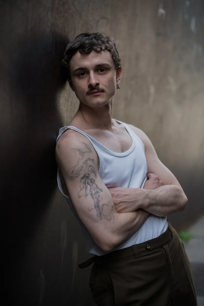 Schauspielerporträt von Felix Steinhilber, der vor einer grauen Betonmauer steht. Er hat die Arme verschränkt und einen Bart. Die Beleuchtung ist gedämpft und erzeugt eine melancholische Stimmung.