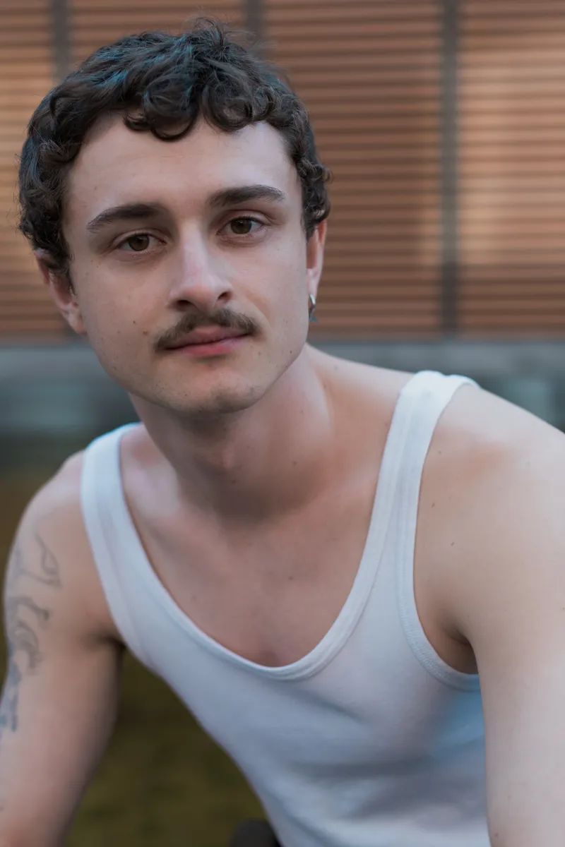 Das Bild zeigt ein Porträt von Felix Steinhilber, einem jungen Schauspieler mit dunklen, gewellten Haaren. Er trägt ein weißes Tanktop und hat eine Tätowierung am Arm. Die Beleuchtung ist weich und warm, was eine entspannte Atmosphäre erzeugt.