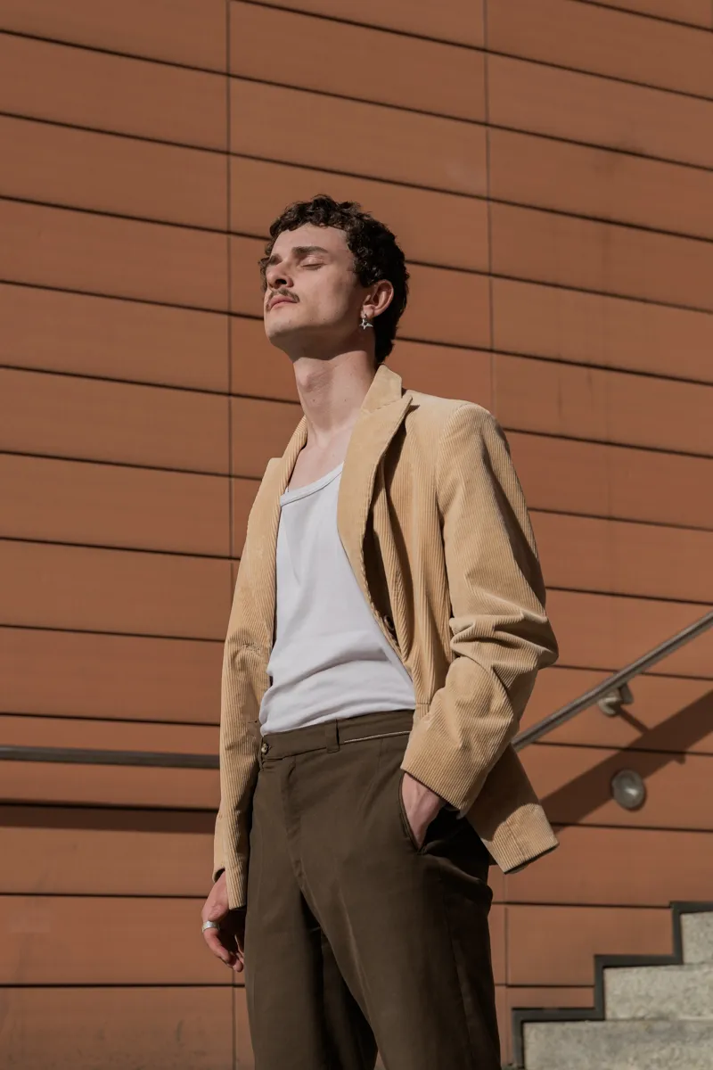 Das Bild zeigt einen jungen Mann, ein Schauspieler, in einem Porträtformat. Er trägt eine Cordjacke, ein weißes T-Shirt und braune Hosen. Die Szene spielt vor einer auffälligen, modernen Fassade mit einer interessanten Fassadenstruktur. Im Hintergrund ist eine Treppe und ein Geländer zu sehen.
