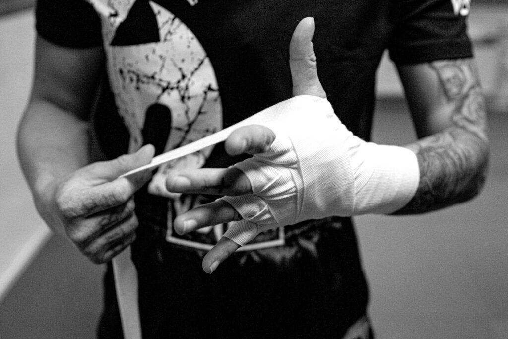 Eine Frau wickelt ihre Hand mit einem weißen Kampfbandage ein. Im Hintergrund ist ein dunkler Raum zu sehen.