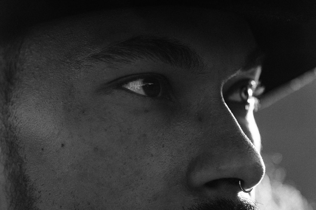 Ein Porträtfoto im Stil eines Seitenprofils, das einen jungen Mann in einer dunklen, stimmungsvollen Aufnahme zeigt. Das Licht fällt seitlich und betont die Konturen des Gesichts, insbesondere das Auge und die Nase. Der Hintergrund ist sehr dunkel oder unscharf, was den Fokus vollständig auf das Subjekt lenkt.