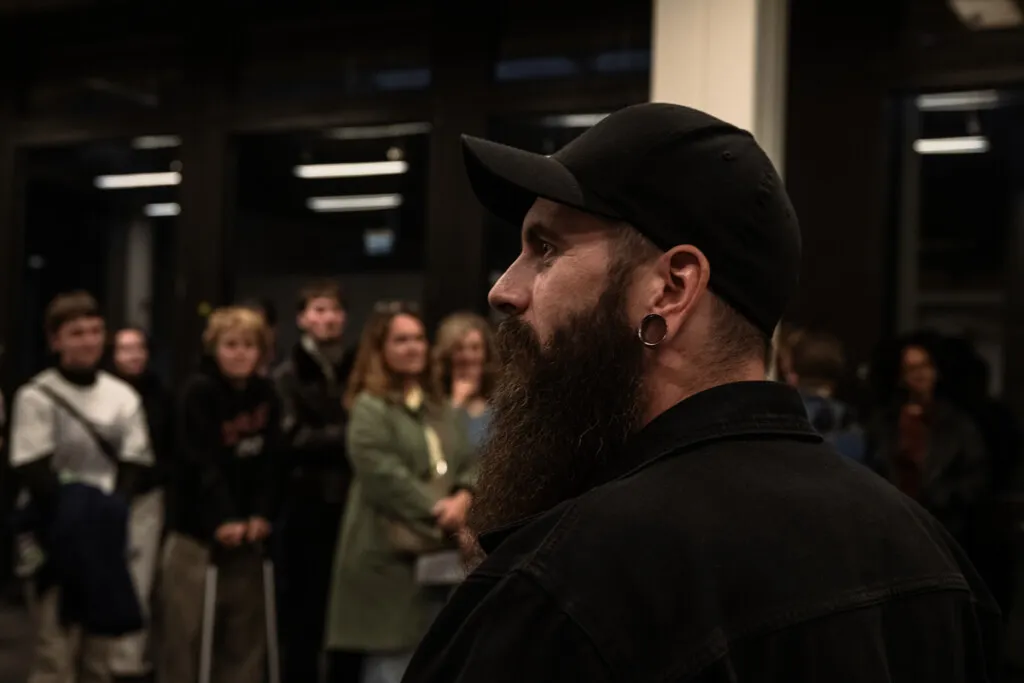 Ein Mann mit Bart und Kopfbedeckung betrachtet eine Gruppe von Menschen, die eine Ausstellung im Zeughof besuchen. Die Szene ist gedimmt und atmosphärisch.