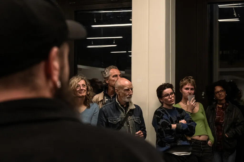 Eine Gruppe von Menschen betrachtet eine Fotografieausstellung des FLUR-Kollektivs im Zeughaus. Der Fokus liegt auf einem Mann im Vordergrund, der mit verschränkten Armen steht. Die Ausstellung ist in einem industriellen Raum mit hohen Decken und Metallverkleidungen stattgefunden.