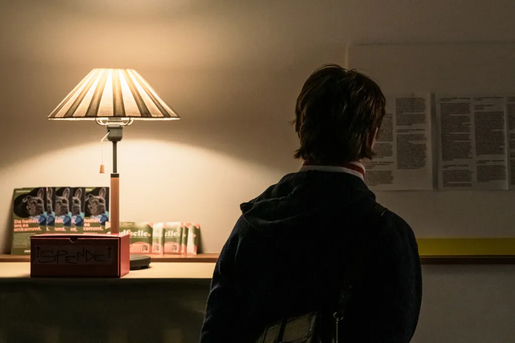 Ein Mann sitzt in einem dunklen Raum, umgeben von Büchern und einer Lampe. Im Hintergrund sind Ausstellungsbeschriftungen und ein Schreibtisch zu sehen. Es ist ein ruhiger, nachdenklicher Moment.