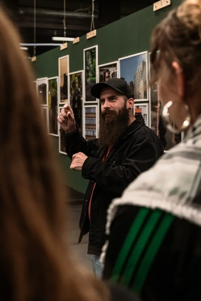 Alexander Friedrich Fotograf in Berlin mit dunkler Mütze erklärt während einer Fotografieausstellung im Zeughof. Im Hintergrund sind Fotografien an der Wand angebracht.