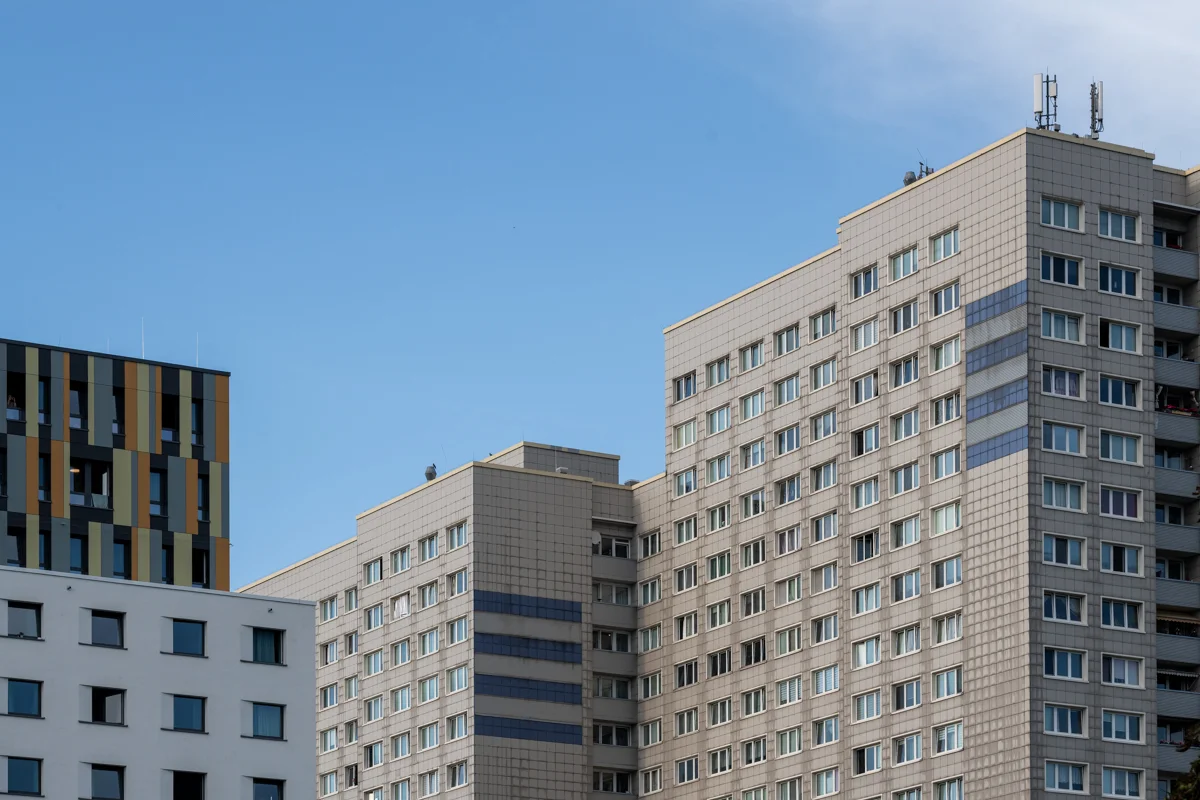Die Aufnahme zeigt eine Reihe von modernen Wohnhäusern, die in einem geometrischen Stil gestaltet sind. Die Gebäude sind aus grauem Ziegelstein oder Beton gefertigt und mit großen Fensterflächen versehen. Der Himmel ist ein klarer, blauer Farbton. Im Vordergrund sind einige Balkone und Fenster zu erkennen. Im Hintergrund sind Antennen auf den Gebäuden zu sehen.