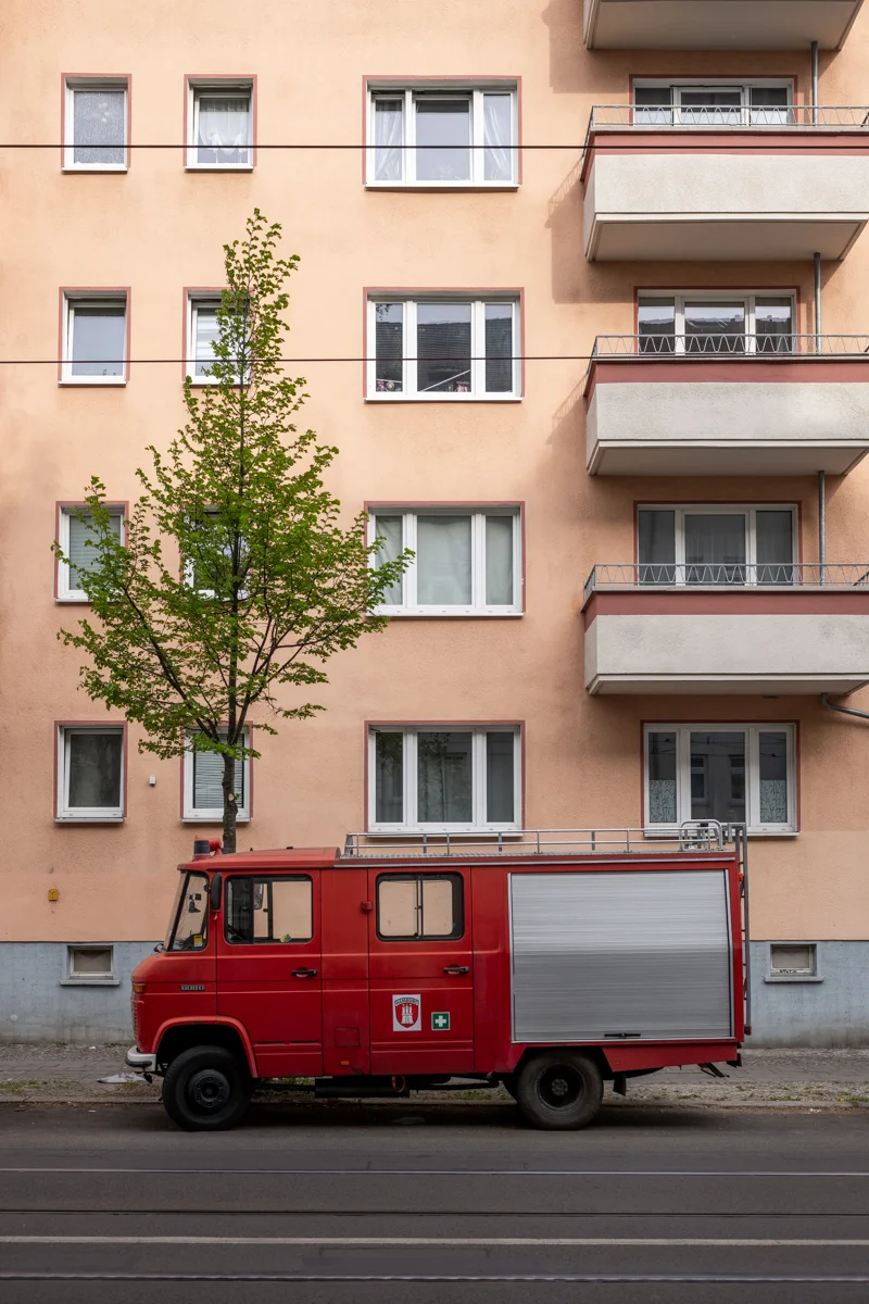 Das Bild zeigt eine straßenseitige Aufnahme eines roten Feuerwehrwagens, vermutlich ein Mercedes-Benz Unimog, der vor einem mehrgeschossigen Wohnhaus steht. Im Vordergrund steht ein Baum mit frischem, jungem Laub. Die Fassade des Wohnhauses ist in einem hellen, rosafarbenen Ton gehalten und mit vielen rechteckigen Fenstern versehen. Die Straße ist mit Stromleitungen versehen. Die Szene wirkt ruhig und urban.