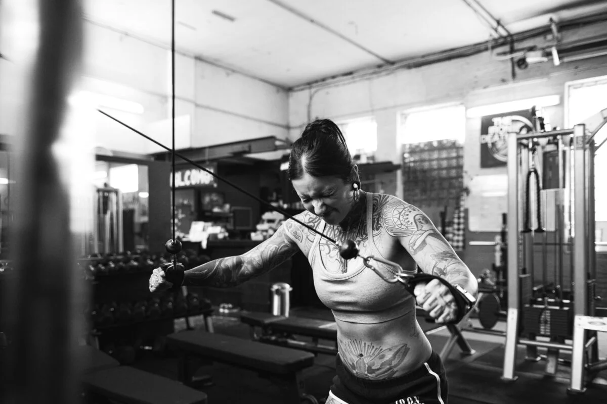 Das Schwarzweißfoto zeigt Personal Trainerin Caro, während sie in einem Fitnessstudio mit einem Kabelzug trainiert. Die Szene ist dynamisch und fokussiert auf die Anstrengung der Sportlerin. Sie befindet sich im Berlin Strength Gym.