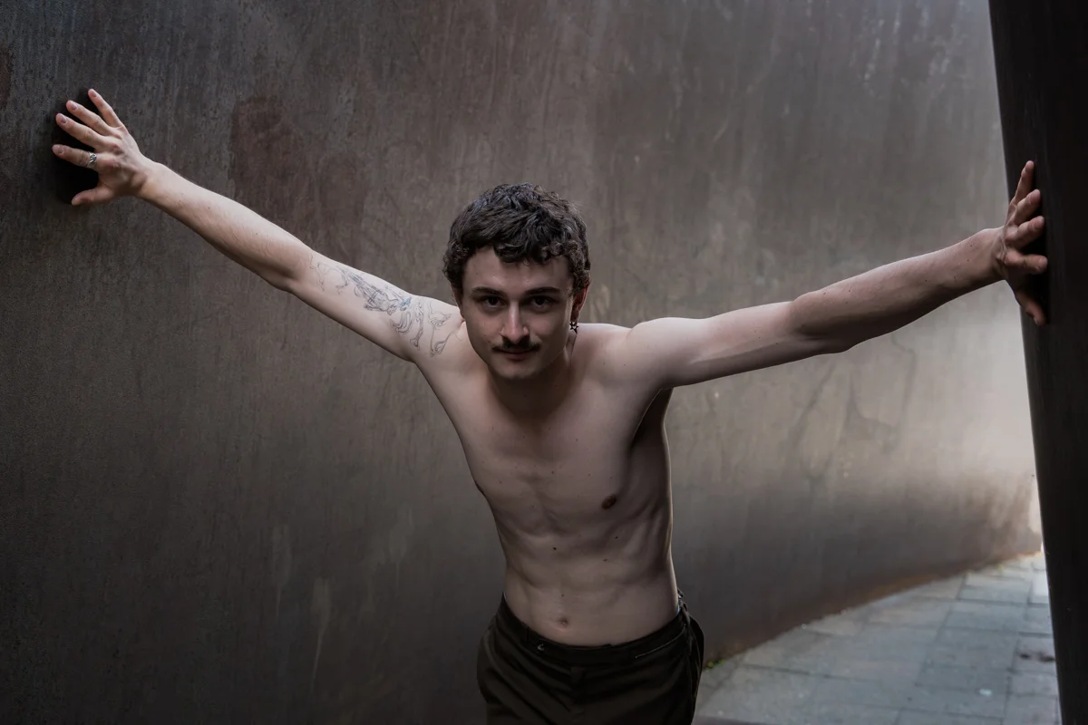 Das Bild zeigt ein Portrait eines Schauspielers, Felix Steinhilber, der vor einer grauen Metallwand mit ausgestreckten Armen steht. Die Pose vermittelt Kontemplation, Verletzlichkeit und eine gewisse Kraft. Der Hintergrund ist dunkel und unscharf, was die Aufmerksamkeit auf den Schauspieler lenkt.