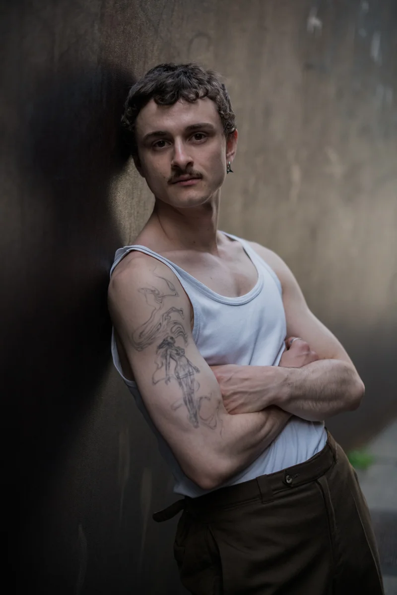 Schauspielerporträt von Felix Steinhilber, der vor einer grauen Betonmauer steht. Er hat die Arme verschränkt und einen Bart. Die Beleuchtung ist gedämpft und erzeugt eine melancholische Stimmung.